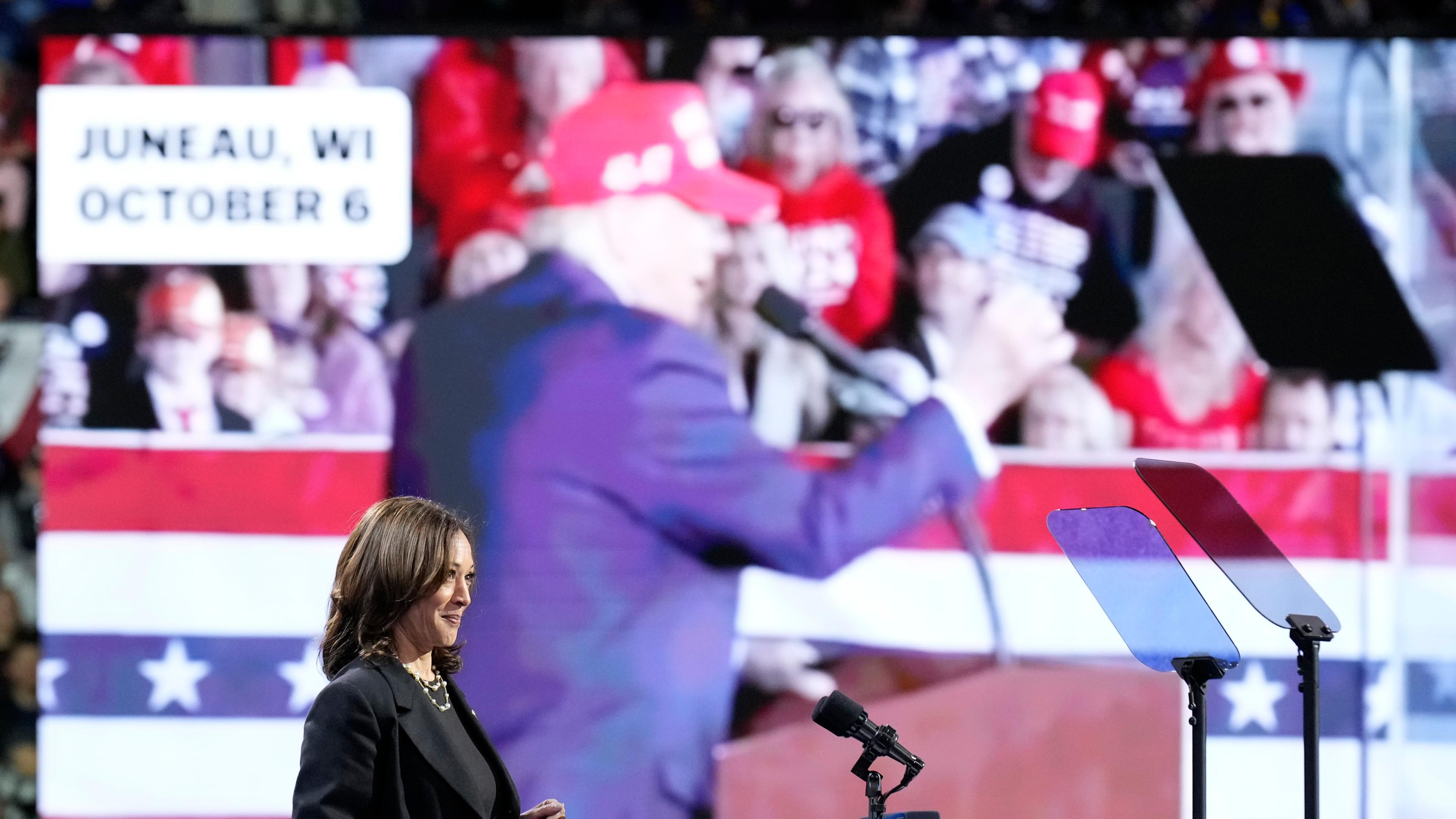 Harris bei Wahlkampfauftritt, auf dem Screen im Hintergrund ist Trump zu sehen.