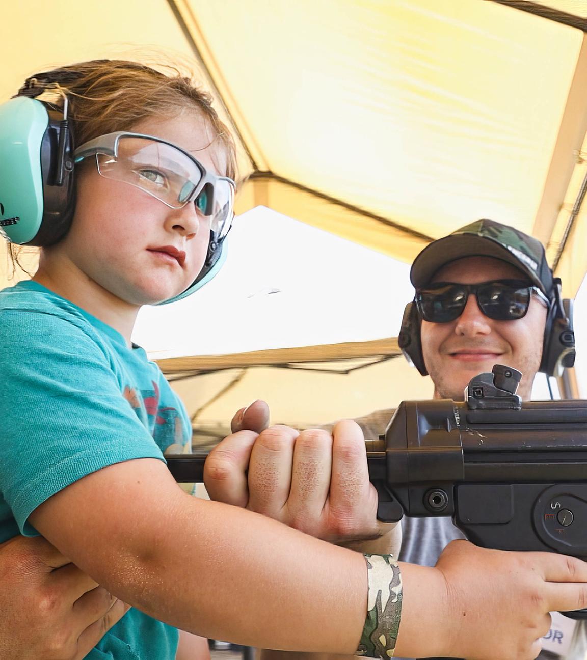 Ein kleines Mädchen mit Ohrschützern hält ein Schnellfeuergewehr. Der Vater trägt eine Sonnenbrille und steht daneben, hält ebenfalls das Gewehr fest und lächelt.