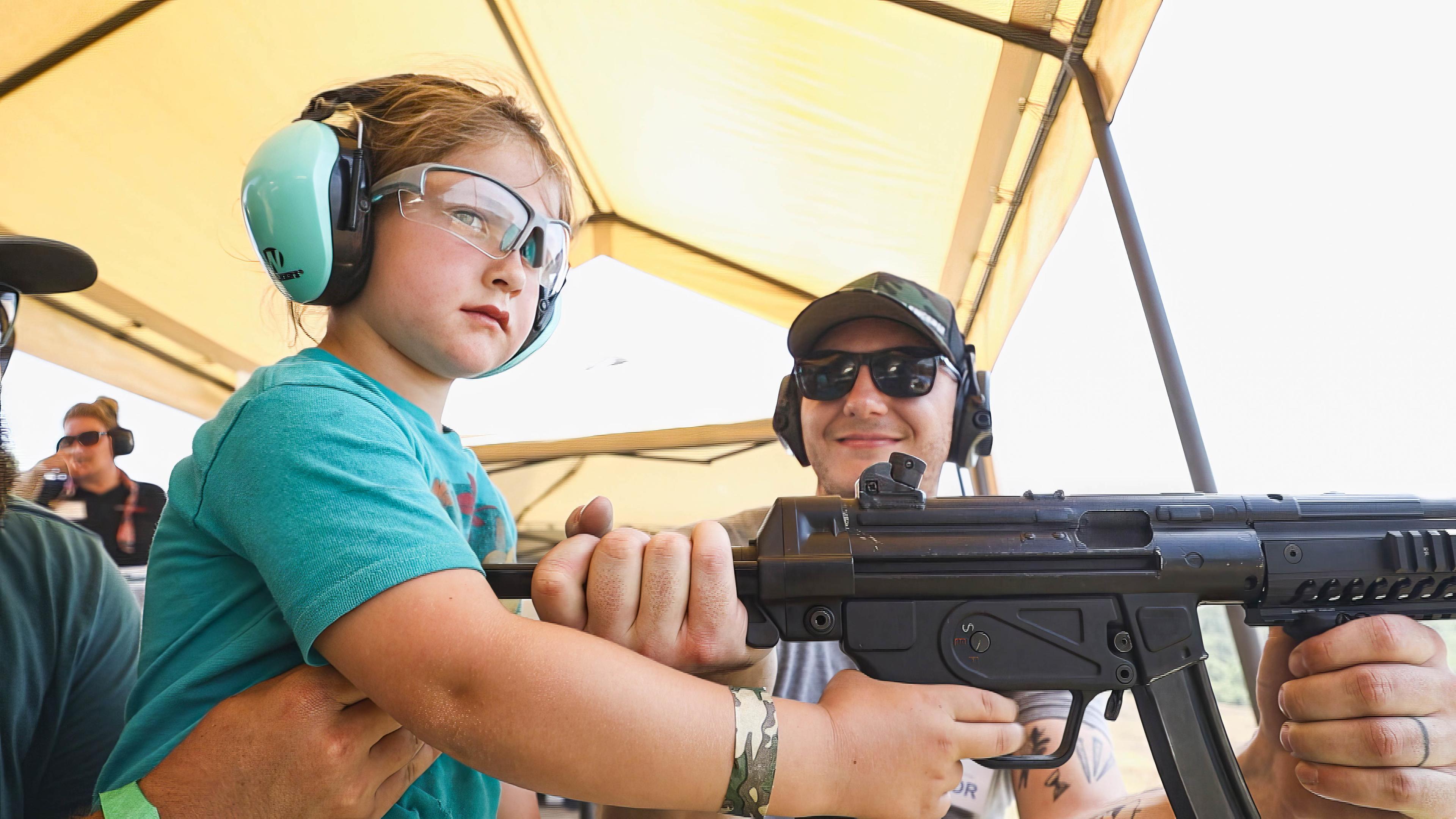 Ein kleines Mädchen mit Ohrschützern hält ein Schnellfeuergewehr. Der Vater trägt eine Sonnenbrille und steht daneben, hält ebenfalls das Gewehr fest und lächelt.