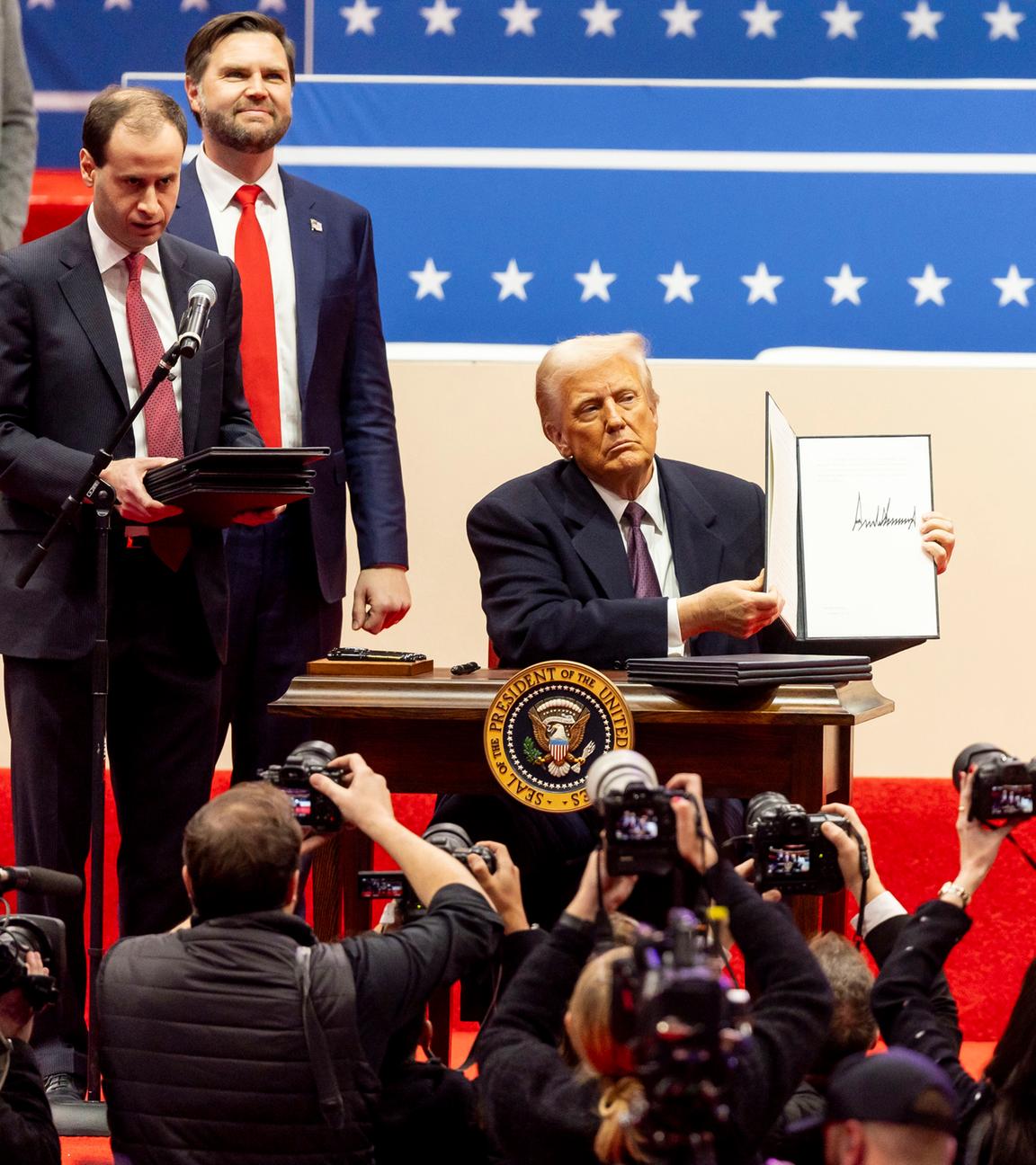 Präsident Trump sitzt auf einer Bühne an einem Schreibtisch. Unter der Bühne stehen Fotografen, hinter ihm steht Vizepräsident J.D. Vance.