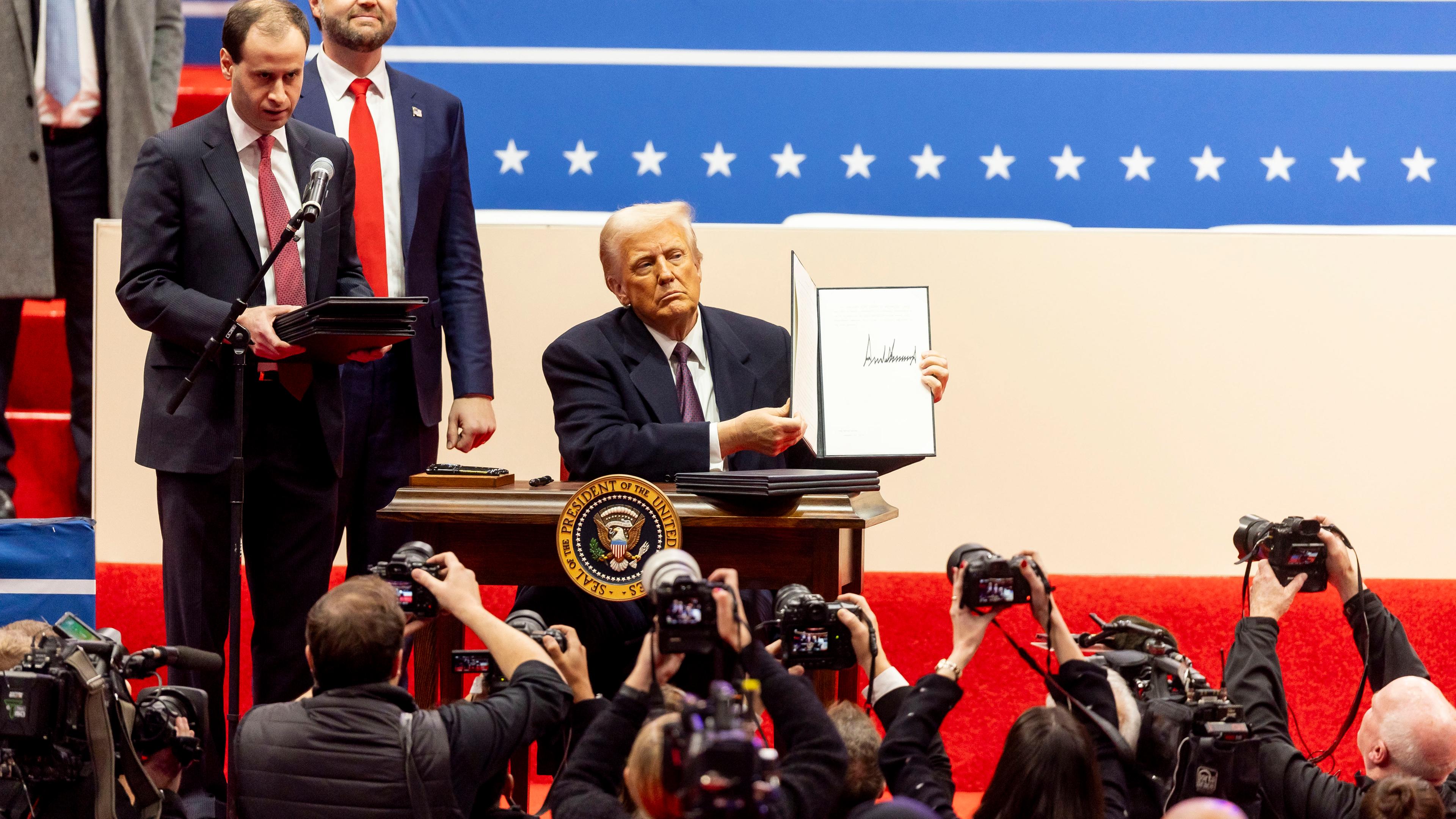 Präsident Trump sitzt auf einer Bühne an einem Schreibtisch. Unter der Bühne stehen Fotografen, hinter ihm steht Vizepräsident J.D. Vance.