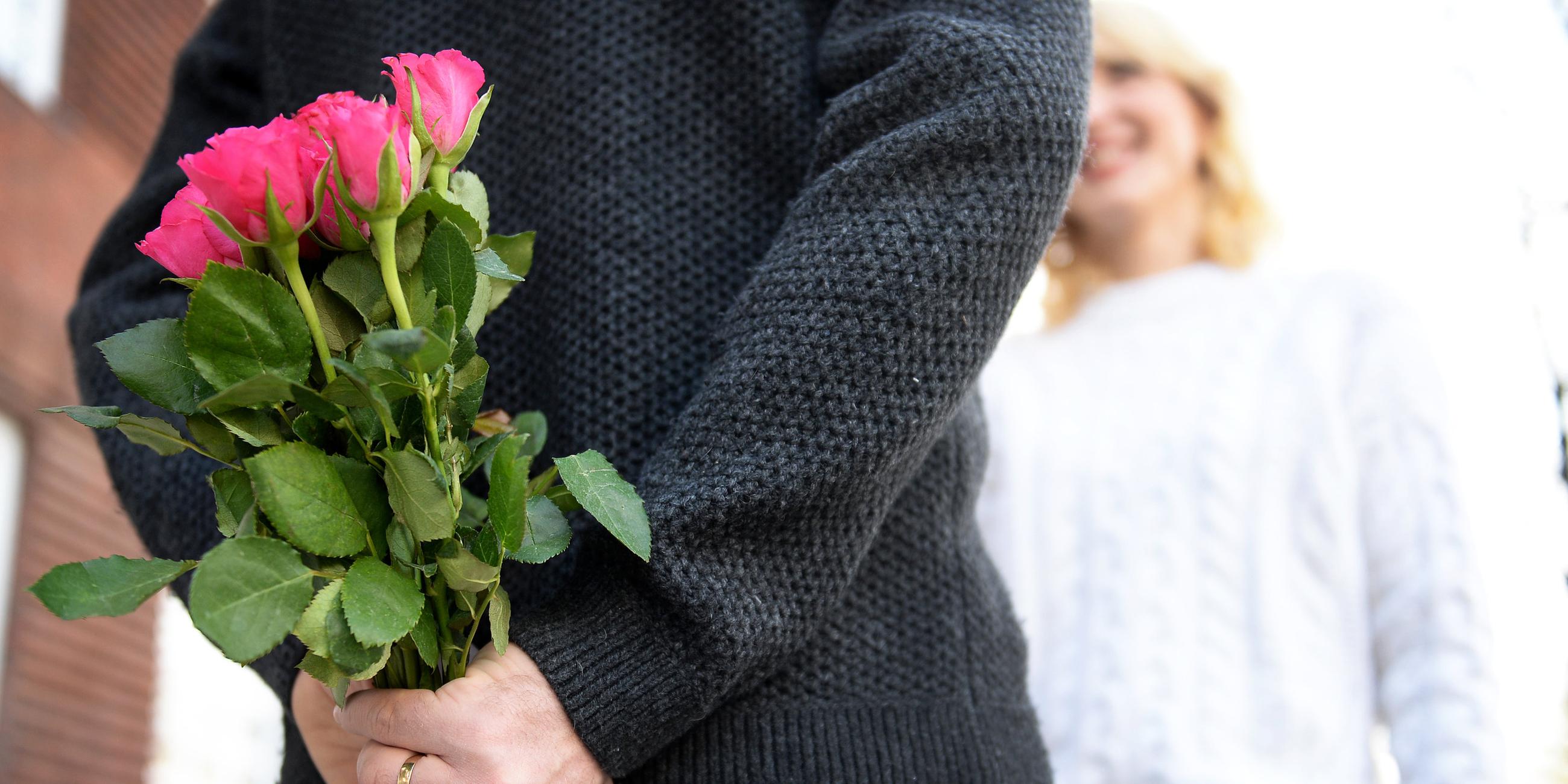 Ein Mann hält einen Strauß Rosen hinter seinem Rücken während eine Frau vor ihm steht.