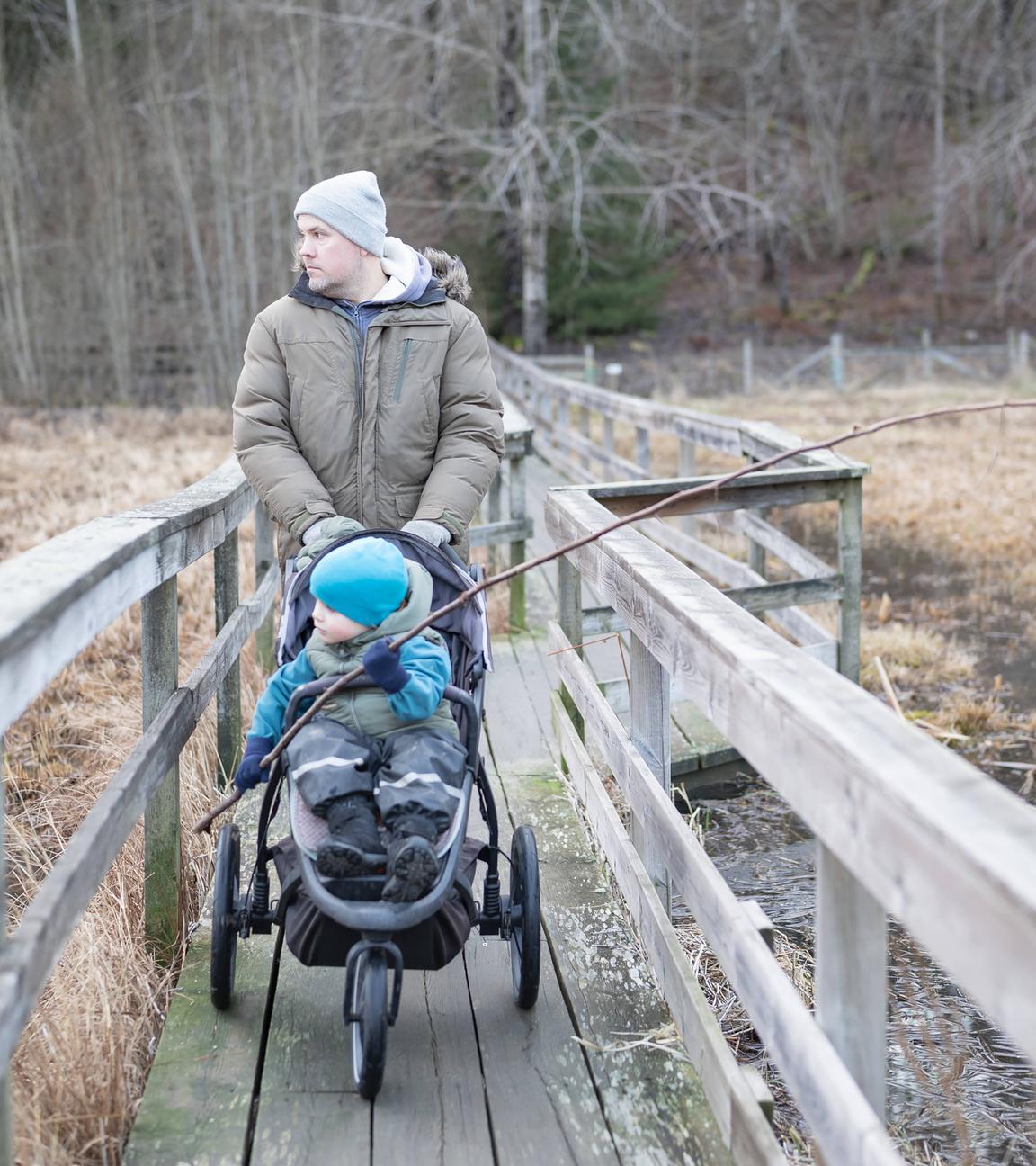 Ein Vater geht mit seinem Kind im Kinderwagen über eine Holzbrücke in der Natur spazieren.