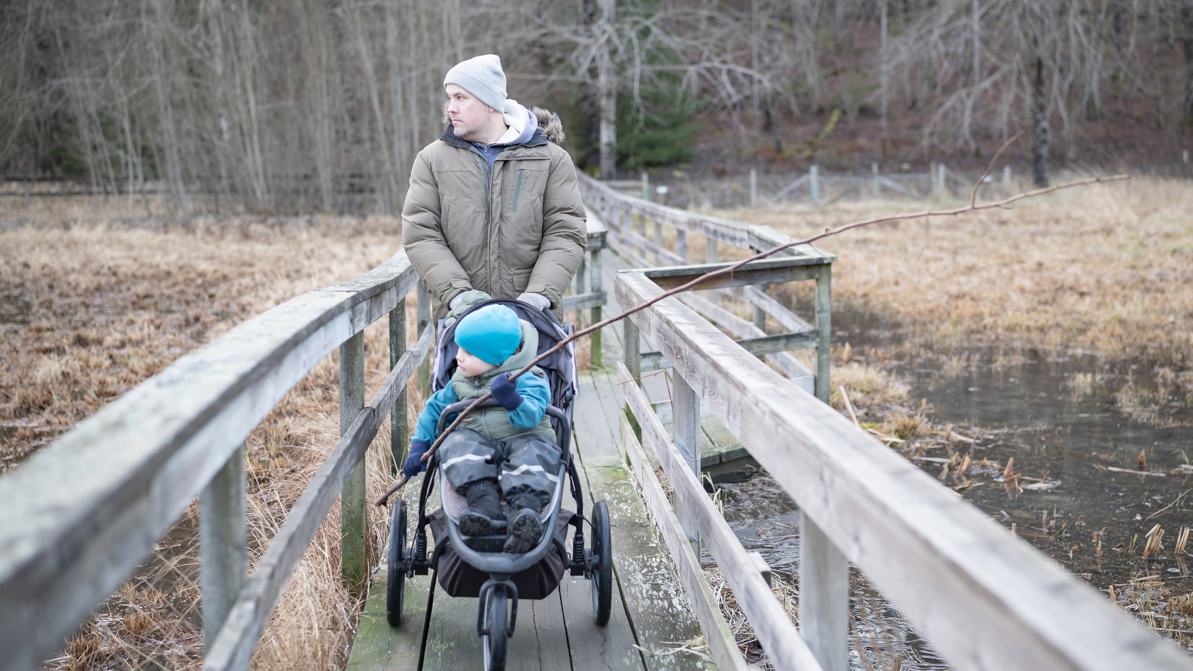 Ein Vater geht mit seinem Kind im Kinderwagen über eine Holzbrücke in der Natur spazieren.