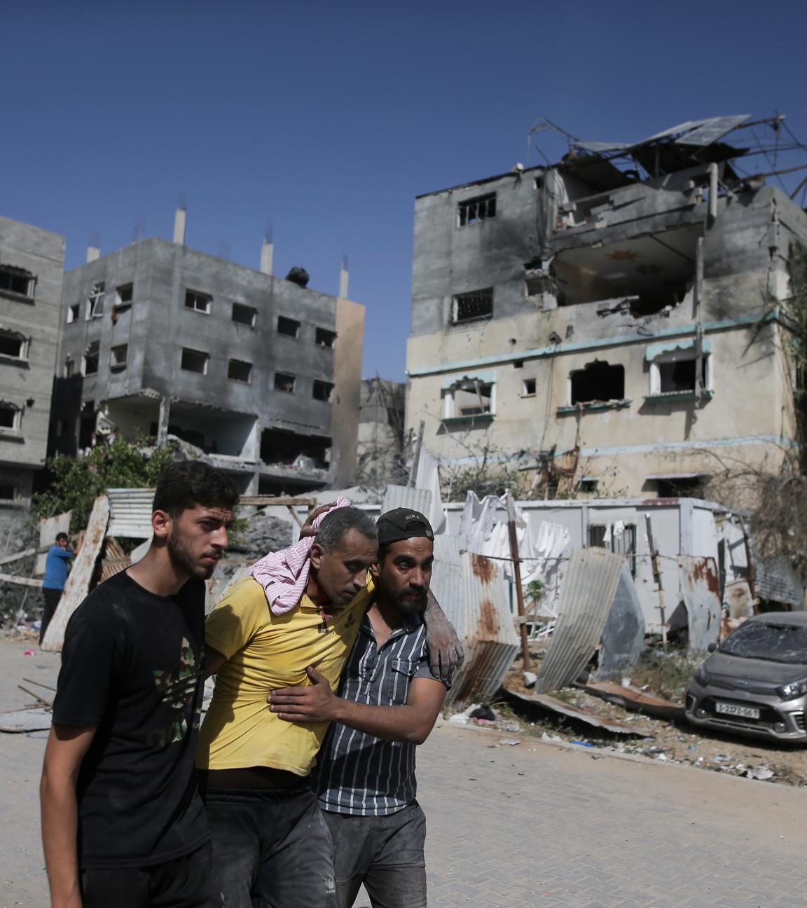 In Nuseirat stützen zwei Palästinenser einen verletzten Mann nach israelischen Angriffen, im Hintergrund sind zerstörte Gebäude zu sehen.