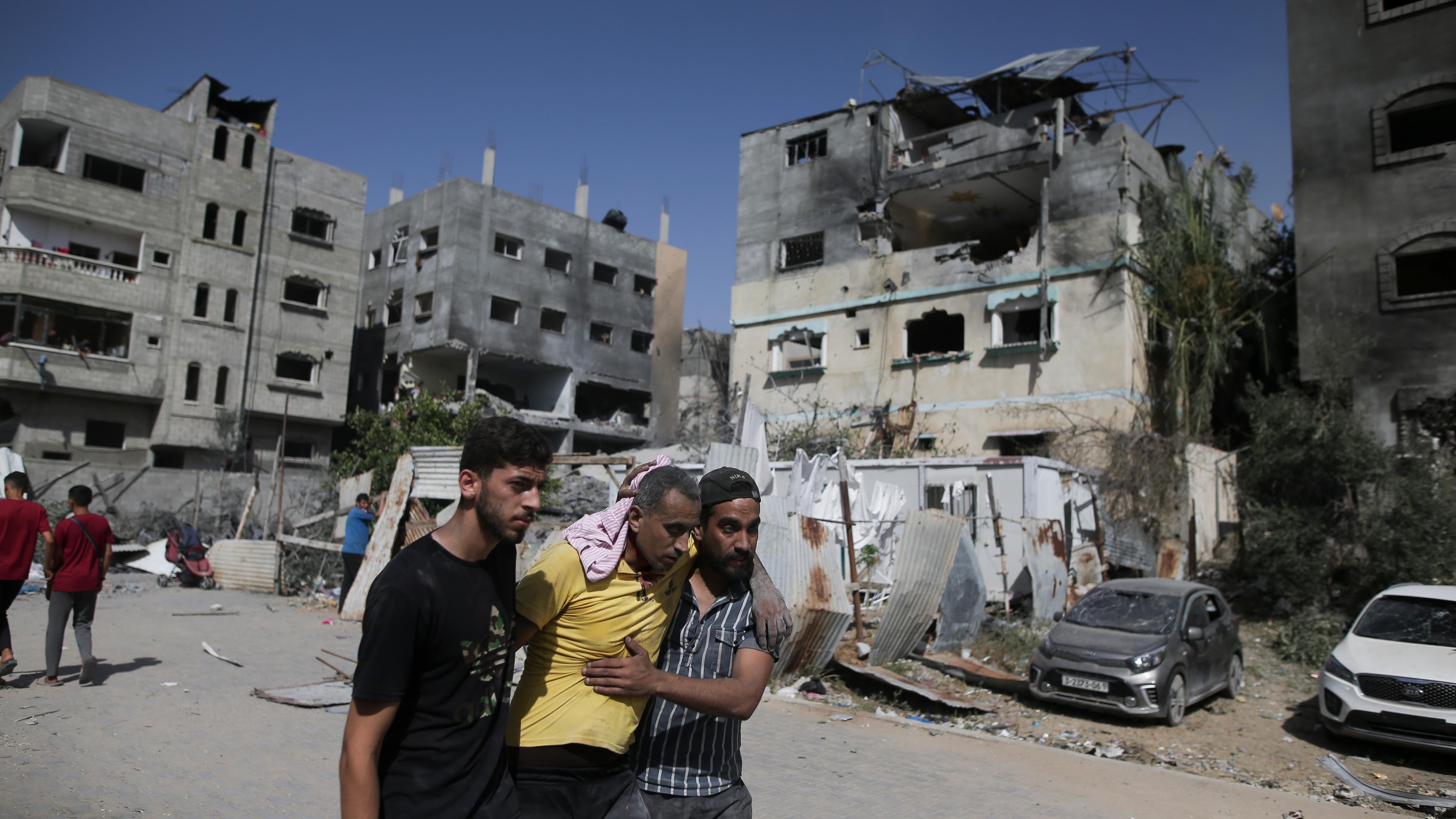 In Nuseirat stützen zwei Palästinenser einen verletzten Mann nach israelischen Angriffen, im Hintergrund sind zerstörte Gebäude zu sehen.