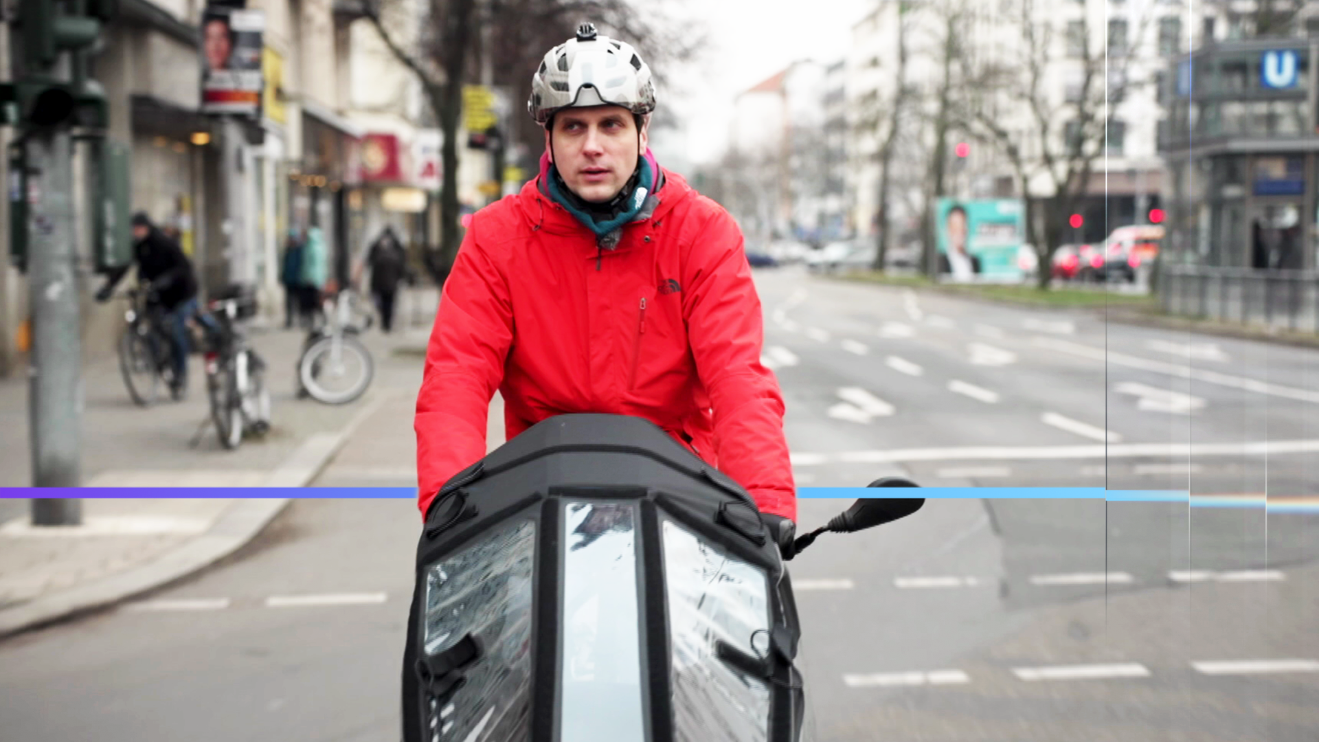 Mann fährt mit Lastenfahrrad eine Straße entlang. Er trägt eine rote Jacke und einen Helm.