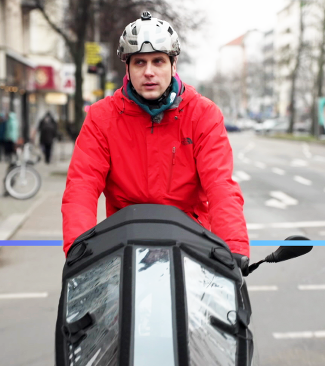 Mann fährt mit Lastenfahrrad eine Straße entlang. Er trägt eine rote Jacke und einen Helm.