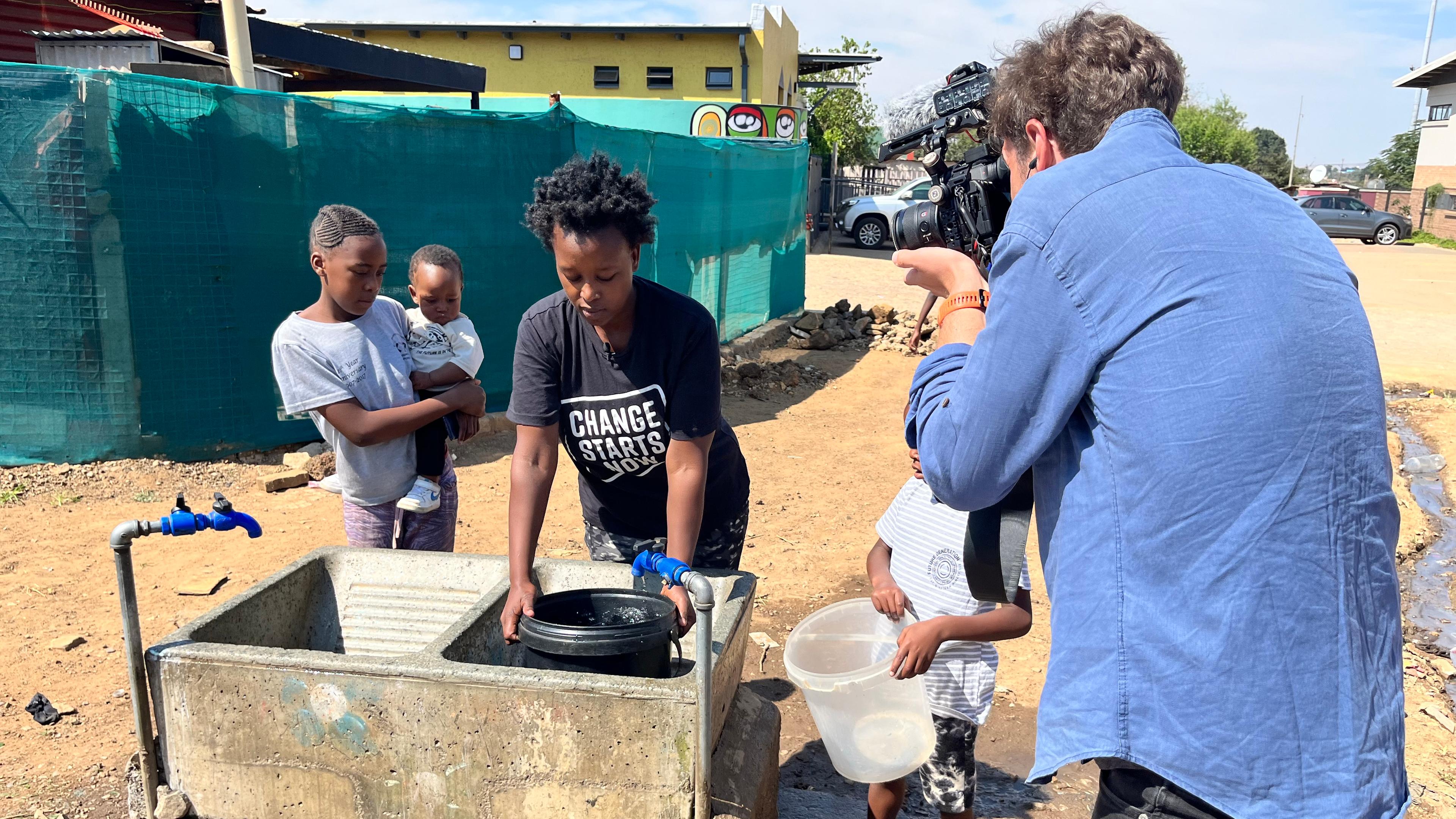 Die junge Süfafrikanerin Lerato Mokhine und ihre Familie befüllen Eimer an einer Wasserstelle in Kliptown, ein Reporter filmt sie dabei.