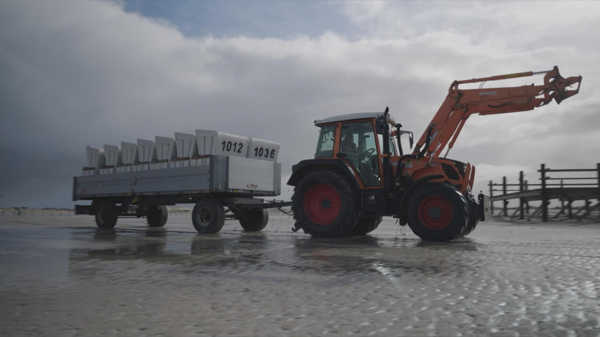 Auf dem Bild ist ein Traktor zu sehen, welcher Strandkörbe über den Strand transportiert.