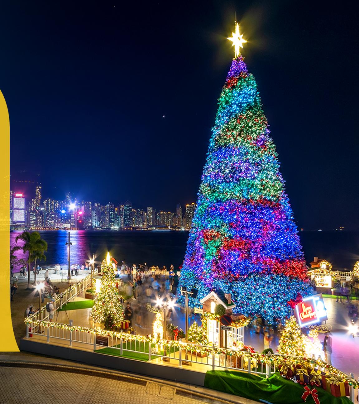 Bunt beleuchteter großer Weihnachtsbaum und Weihnachtsdeko am Hafen von Hongkong bei Nacht, im Hintergrund beleuchtete Skyline der Stadt, daneben ein großes gelbes a