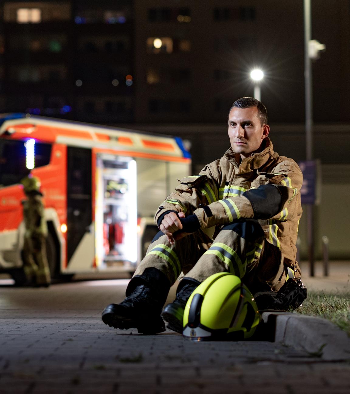 Feuerwehrmann Adrian sitzt in Feuerwehrmontur auf dem Bordstein, im Hintergrund ein Feuerwehrauto.