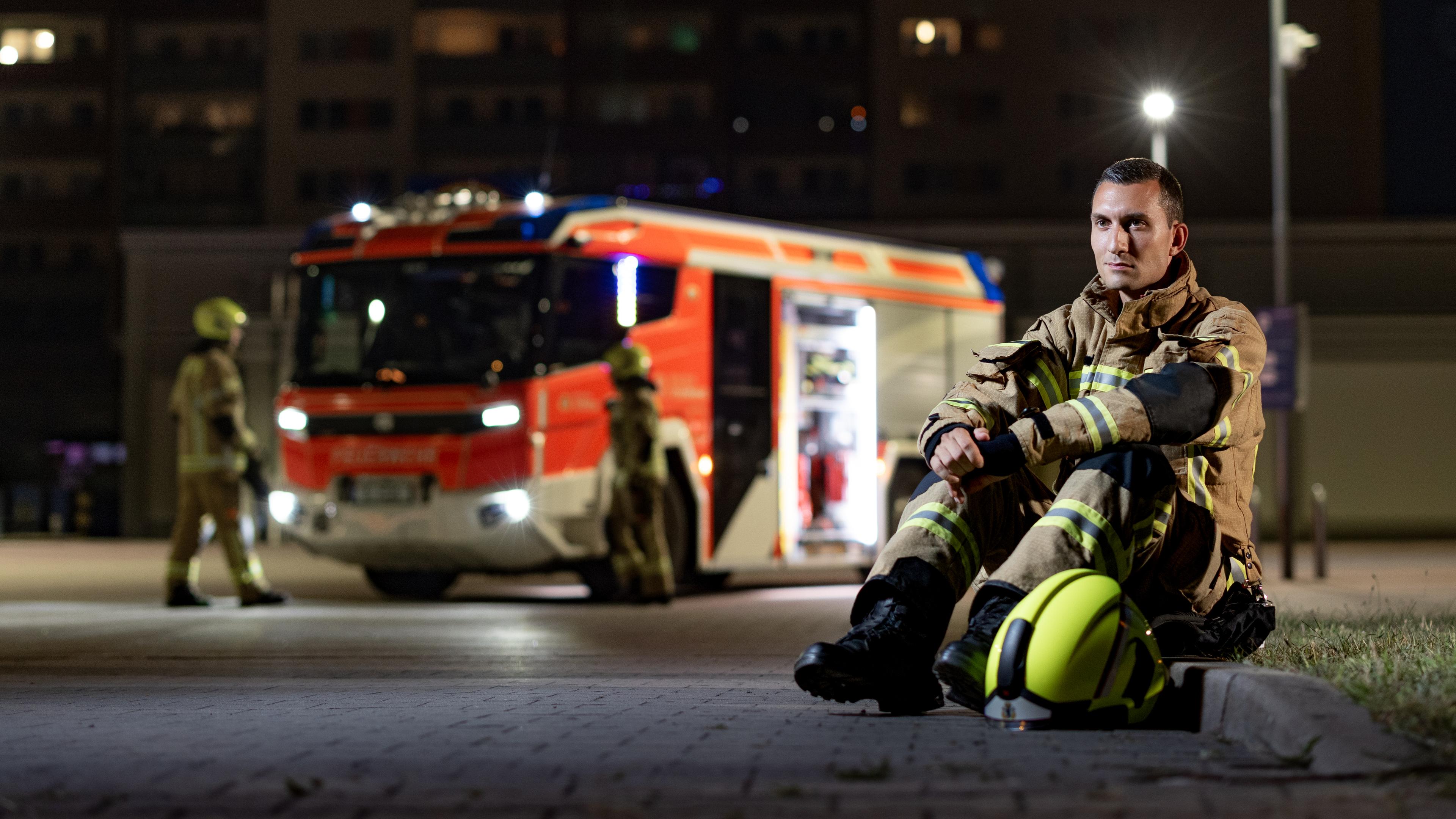 Feuerwehrmann Adrian sitzt in Feuerwehrmontur auf dem Bordstein, im Hintergrund ein Feuerwehrauto.