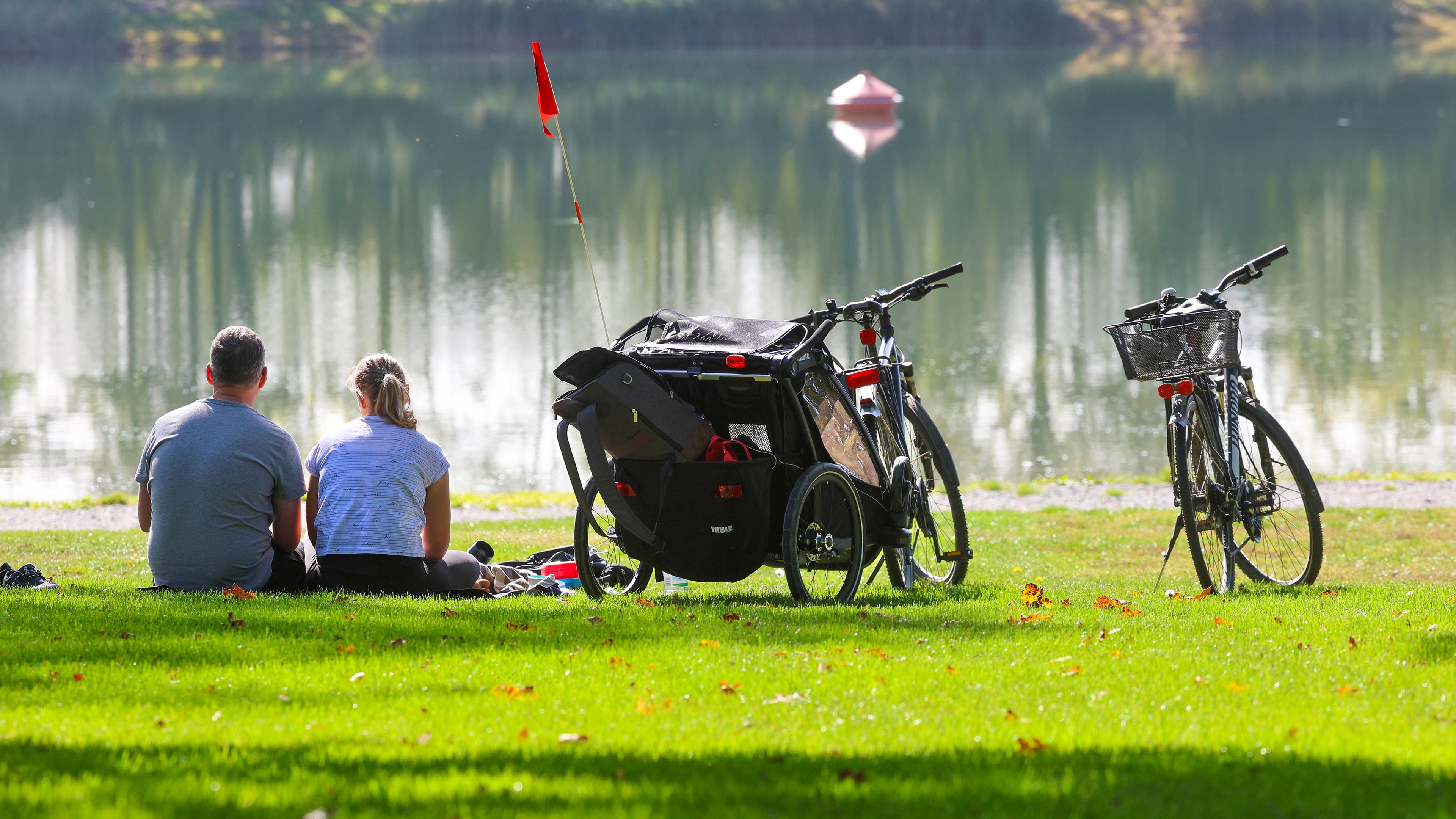 Baden-Württemberg, Ertingen: Ein Mann und eine Frau sitzen neben ihren Fahrrädern an einem See in der Sonne