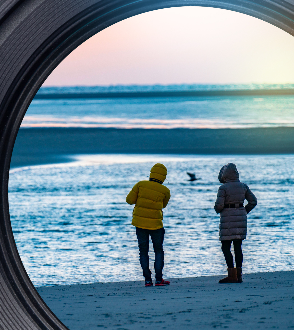 Zwei Menschen in Winterjacken am Strand bei Sonnenuntergang.