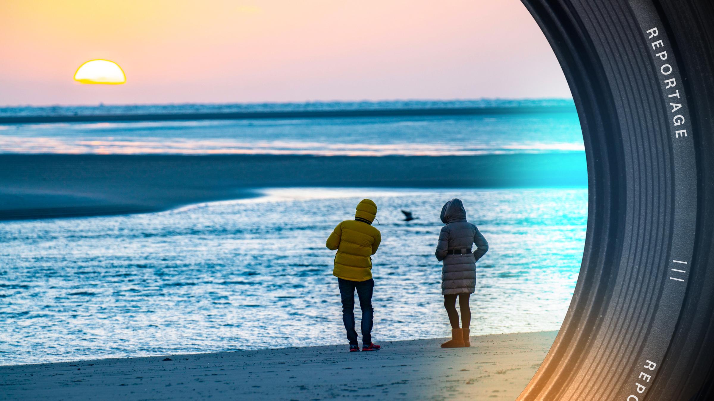 Zwei Menschen in Winterjacken am Strand bei Sonnenuntergang.