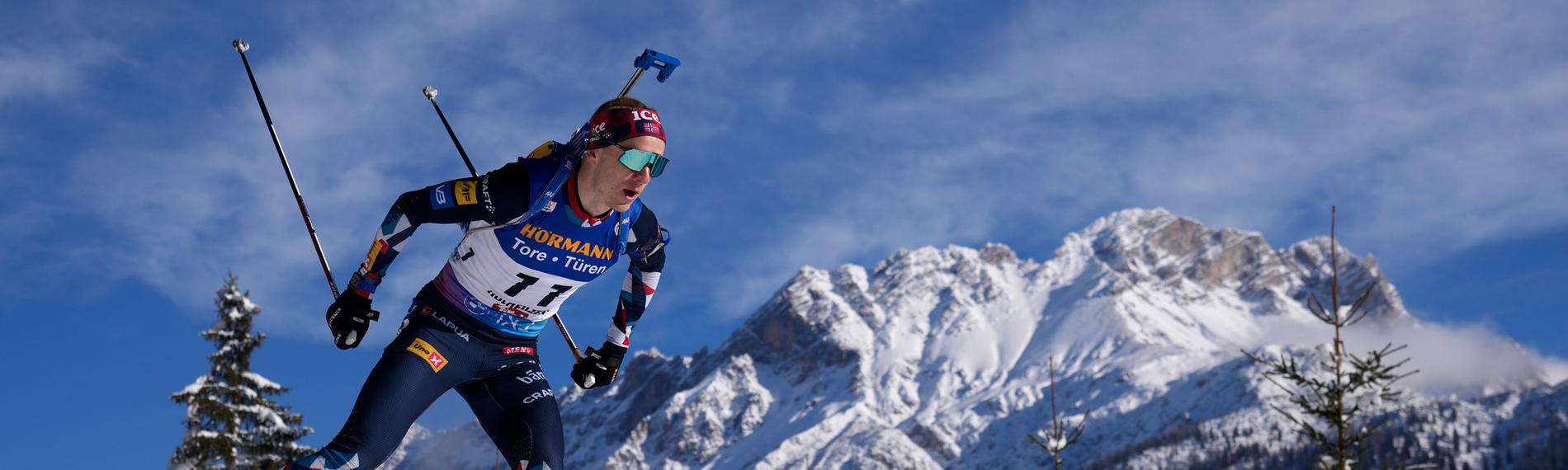 Ein Biathlon-Sportler auf einer Piste mit verschneiten Bergen im Hintergrund.