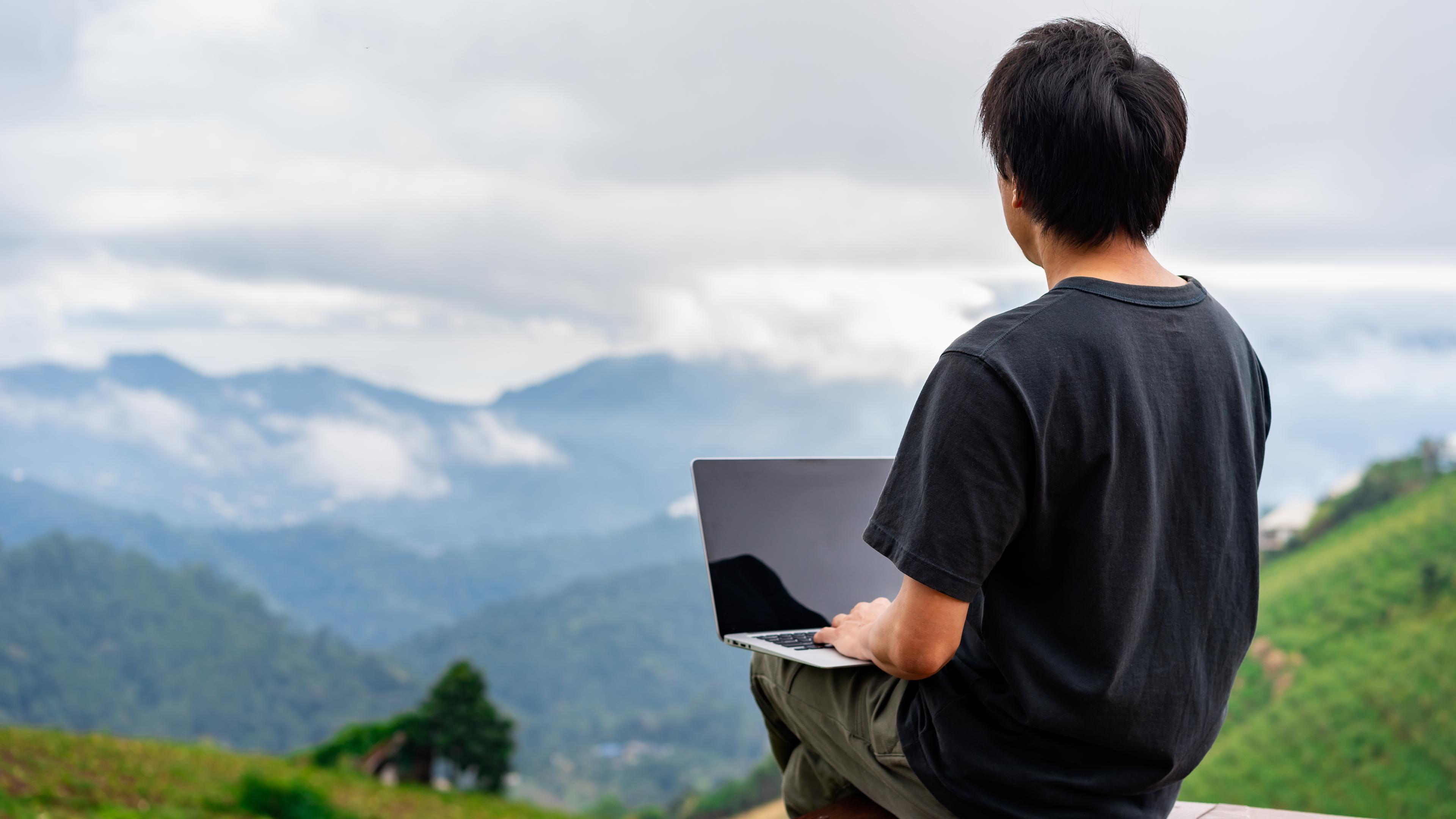 Junger Mann sitzt mit Laptop auf einer Holzfläche und blickt in die Berge.