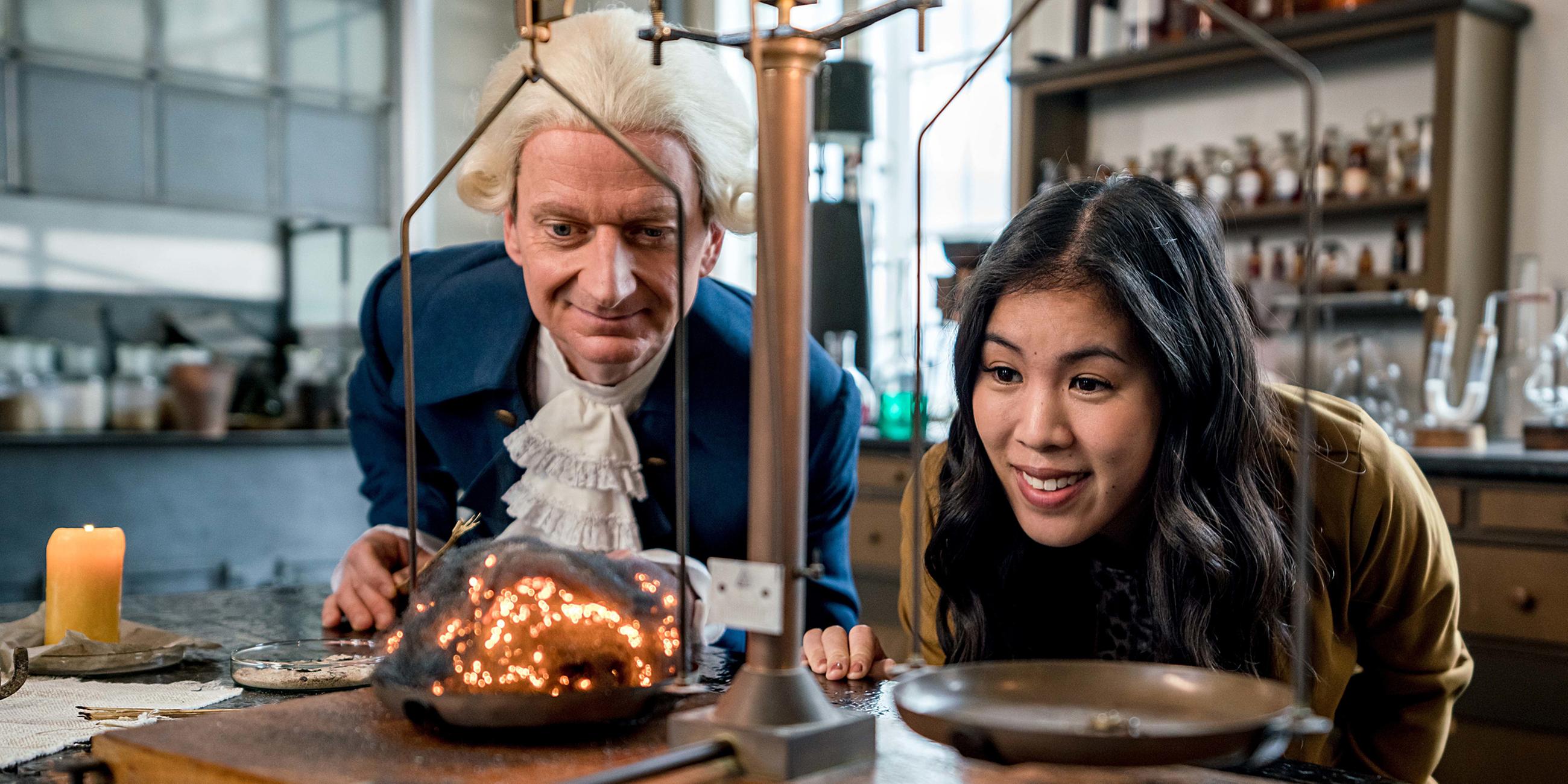 Mai Thi Nguyen-Kim und Michael Kessler (als Darsteller des Wissenschaftlers Joseph Priestley) blicken in einem Labor auf eine Waage.