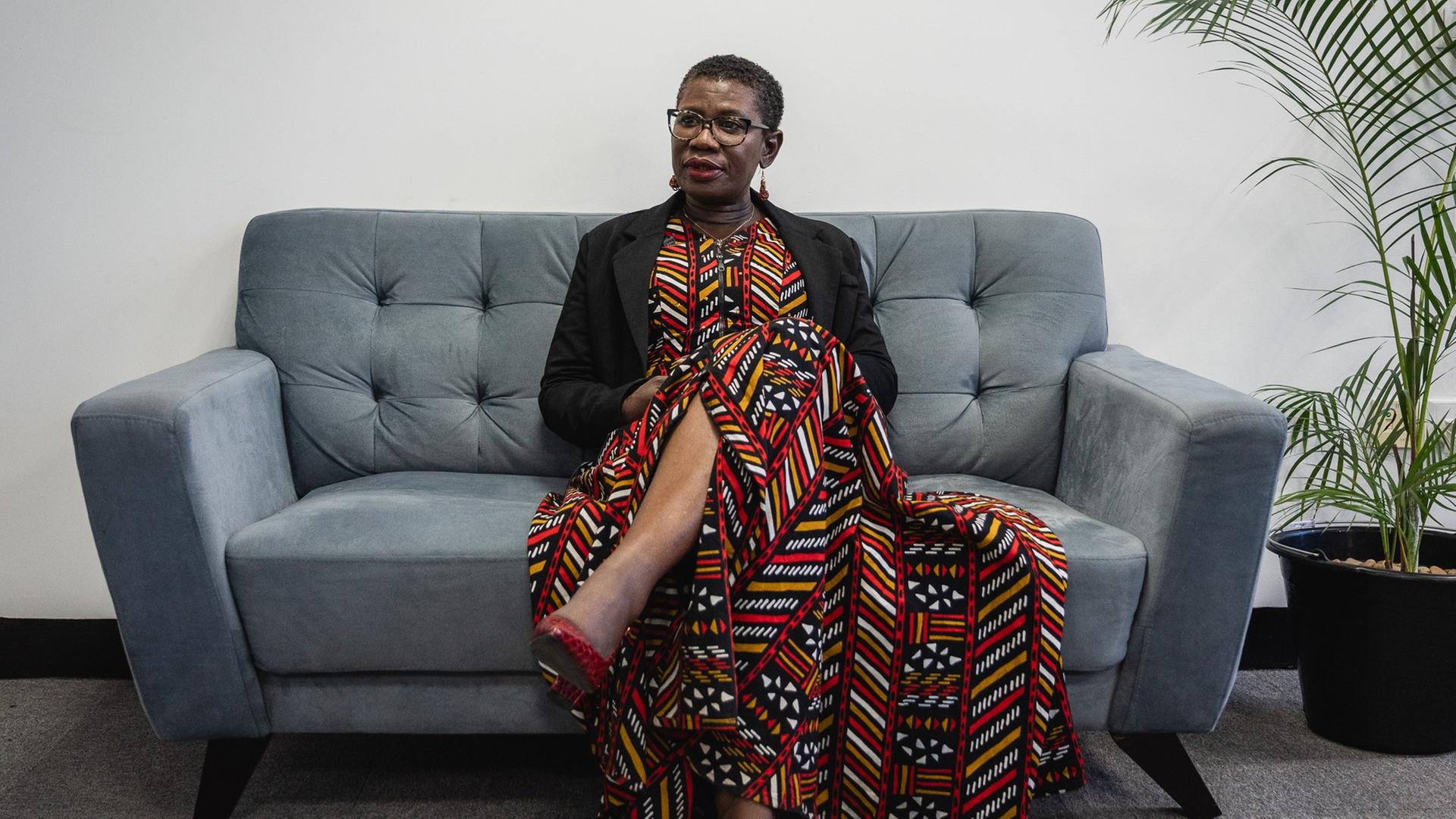 Ein Frau mit kurzen Haaren und einem traditionellen afrikanischen Kleid sitzt auf einem grauen Sofa.