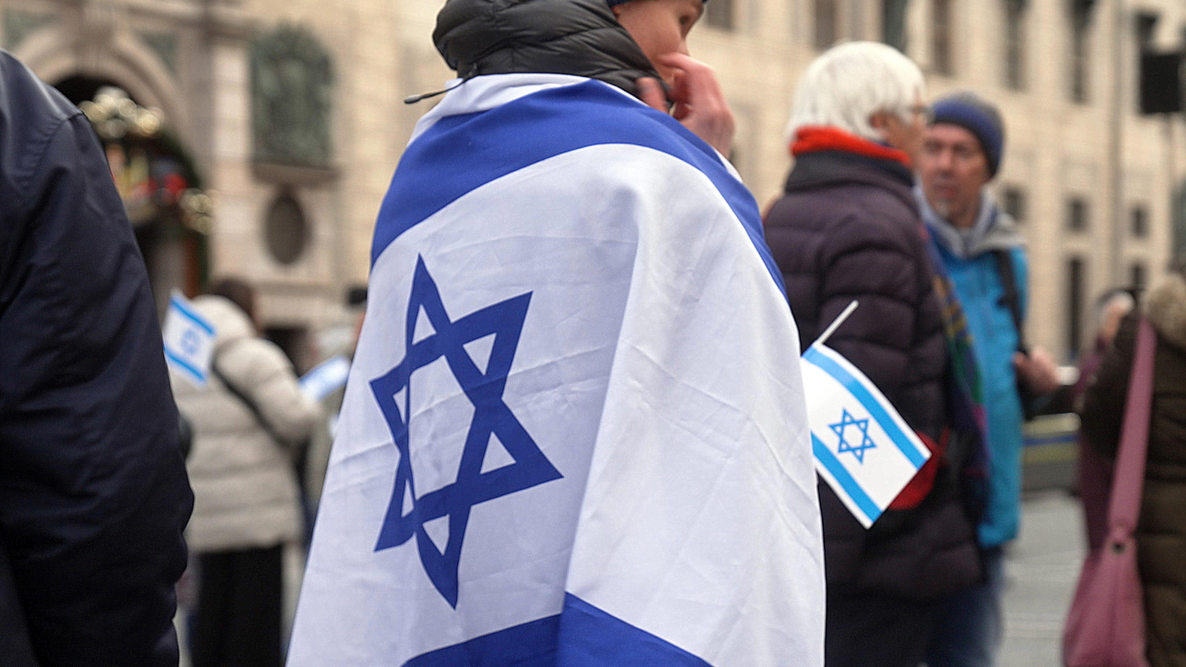 Demo. Frau von hinten in Israel-Flagge gehüllt. Im Hintergrund unscharf weitere Demo-Teilnehmer.